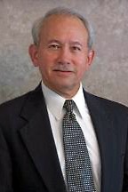 Professional man in a dark suit and white shirt with a patterned tie, smiling against a neutral background. Suitable for business or leadership contexts.